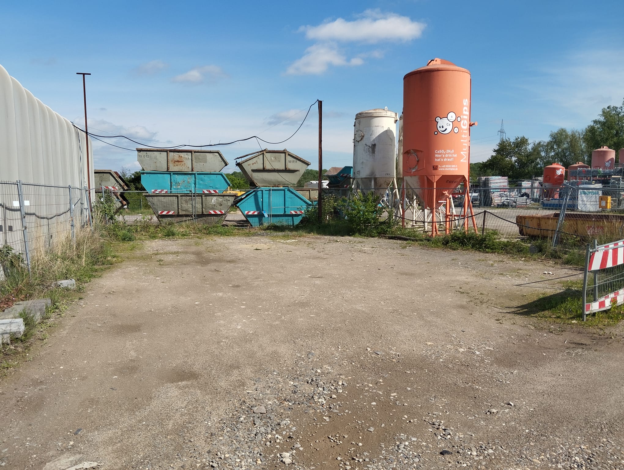 Ein leerer, sonniger Platz mit Containern, Zäunen und einem orangen Silo