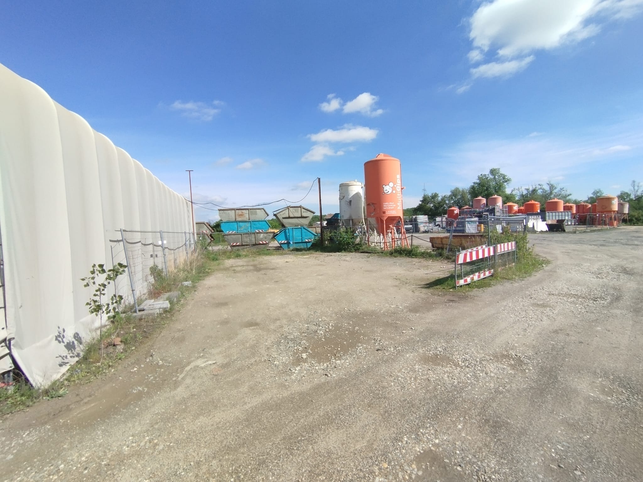 Ein sonniger Industriehof mit orangen Silos, Containern und grünen Büschen