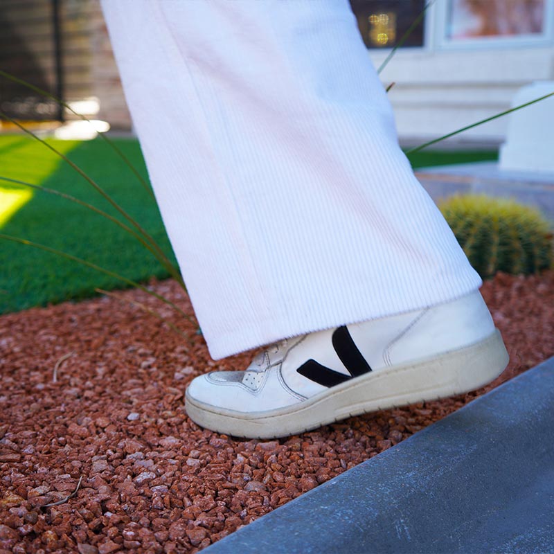 Weisser Sneaker mit schwarzem V Logo unter heller Cordhose auf rotem Kies