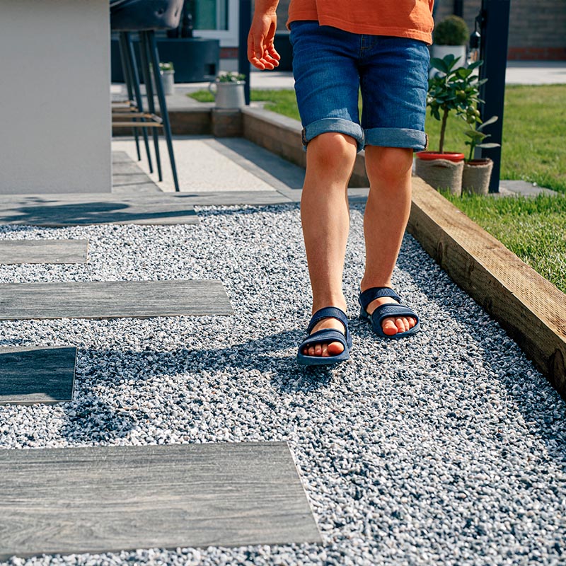Eine Person in Jeansshorts und blauen Sandalen geht ueber einen Kiesweg