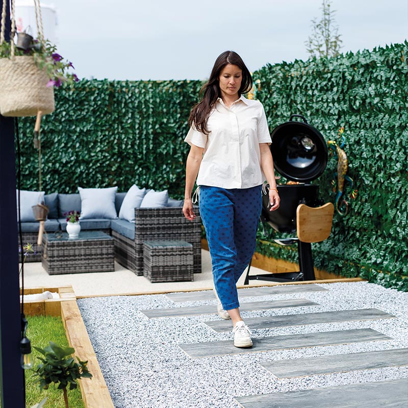 Frau mit weisser Bluse und blauer Hose geht auf einer Terrasse mit Kunsthecke