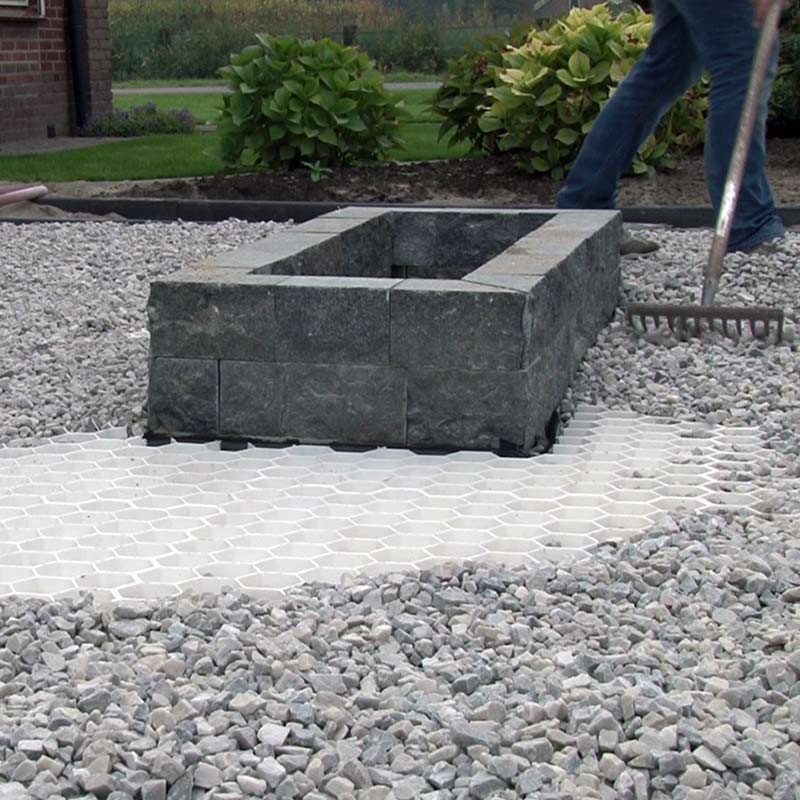 Person in Jeans verteilt grauen Kies mit einem Rechen neben Steinbeet im Garten