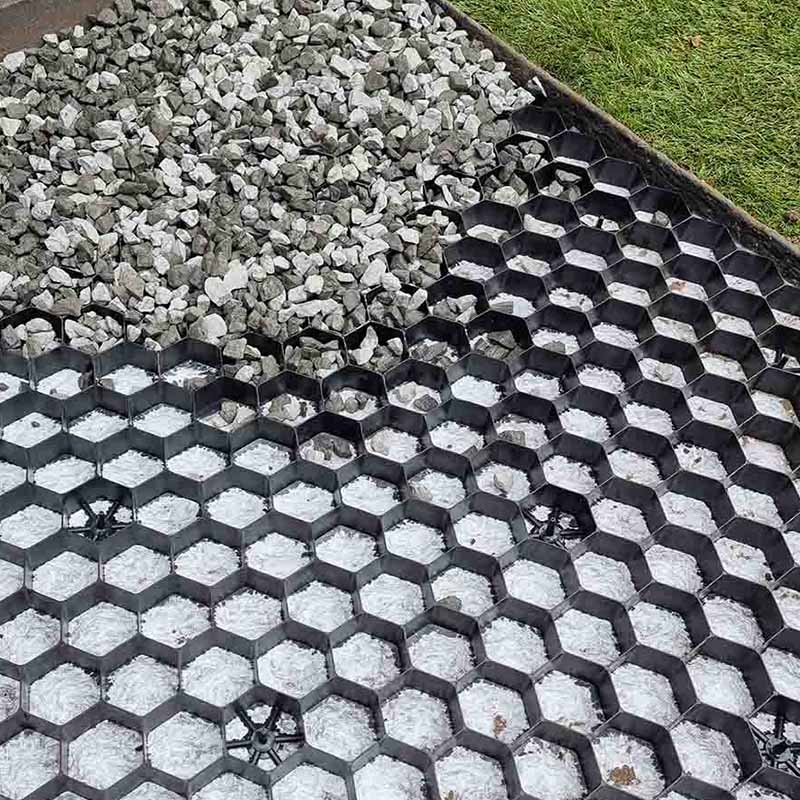 Schwarze Wabengitter aus Kunststoff mit grauem Schotter neben gruenem Rasen
