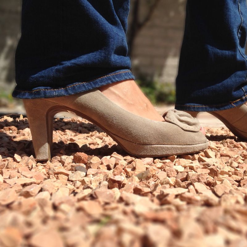 Person in beigen Wildleder Peeptoe High Heels und dunkler Jeans auf Kies